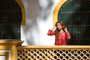 Beautiful young woman in a typical Moroccan red suit, embroidered with gold and silver threads, touching her hair in an outdoor park. Concept beauty, ethnicity, typical suits, Marrakech, arab.
