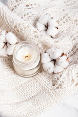 Obraz na płótnie Canvas Scented candle in a glass and cotton flowers on white knitted plaid. Vertical. Burning candle