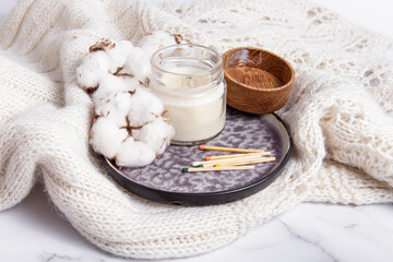 Scented candle in a glass with wooden lid, cotton flowers and wooden matches on white knitted plaid. Mock up. Cozy