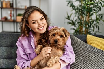 Young beautiful hispanic woman hugging dog sitting on sofa at home