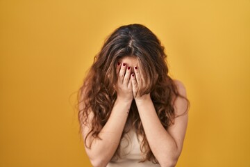Young hispanic woman standing over yellow background with sad expression covering face with hands...