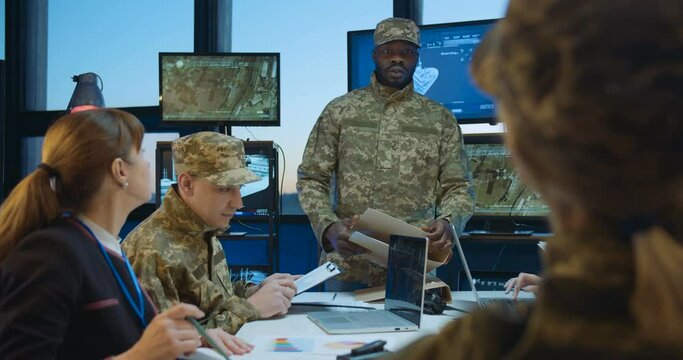 African American young man, head of headquarters coming in and handing documents with tasks to Caucasian troops workers. Metting about army studing and planning. Brainstorming of army operation.