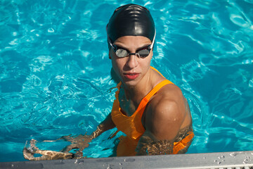 athletic female swimmer in orange swimsuit, swimming cap and goggles standing in swimming pool near border