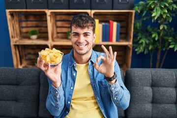 Young hispanic man holding potato chips doing ok sign with fingers, smiling friendly gesturing excellent symbol
