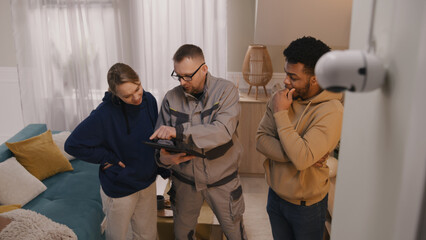 Installer sets up security camera in apartment using digital tablet. Man shows to multi cultural...