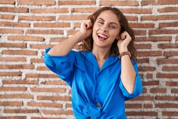 Beautiful brunette woman standing over bricks wall smiling pulling ears with fingers, funny...