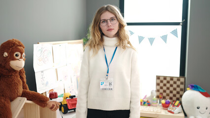 Young blonde woman preschool teacher standing with relaxed expression at kindergarten