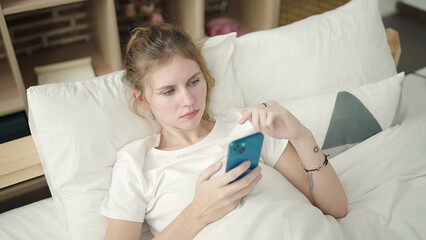 Young blonde woman using smartphone lying on bed at bedroom