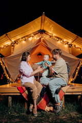 Obraz na płótnie Canvas Happy family with lovely baby relaxing and spend time together in glamping on summer evening near cozy bonfire. Luxury camping tent for outdoor recreation and recreation. Lifestyle concept