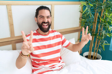 Young hispanic man doing victory gesture with fingers sitting on bed at bedroom