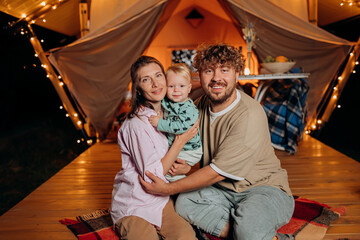 Obraz na płótnie Canvas Happy family with lovely baby relaxing and spend time together in glamping on summer evening near cozy bonfire. Luxury camping tent for outdoor recreation and recreation. Lifestyle concept
