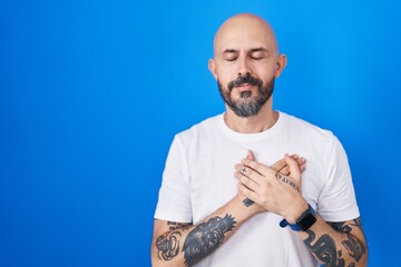 Hispanic man with tattoos standing over blue background smiling with hands on chest with closed eyes and grateful gesture on face. health concept.