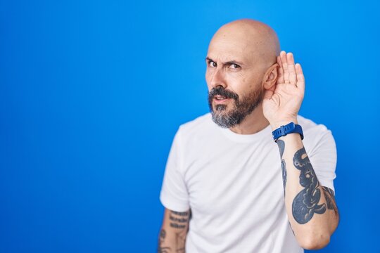 Hispanic man with tattoos standing over blue background smiling with hand over ear listening an hearing to rumor or gossip. deafness concept.