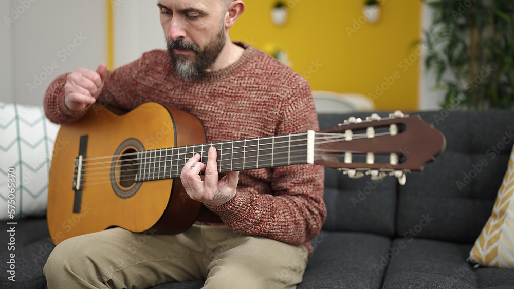 Sticker Young bald man playing classical guitar sitting on sofa at home