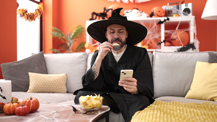 Young bald man wearing wizard costume eating chips potatoes using smartphone at home