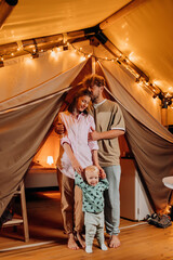 Obraz na płótnie Canvas Happy family with lovely baby relaxing and spend time together in glamping on summer evening. Luxury camping tent for outdoor recreation and recreation. Lifestyle concept