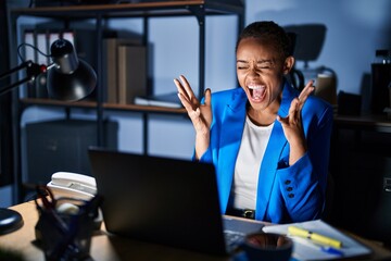 Beautiful african american woman working at the office at night celebrating mad and crazy for success with arms raised and closed eyes screaming excited. winner concept