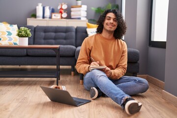 Obraz premium Young latin man using laptop writing on notebook at home