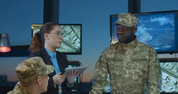 Multiethnic staff of militarian headquarters having urgent meeting on trooops dislocation in office. Female assistant coming and bringing new data and information in documents.
