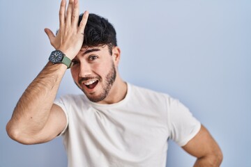 Hispanic man with beard standing over white background surprised with hand on head for mistake,...