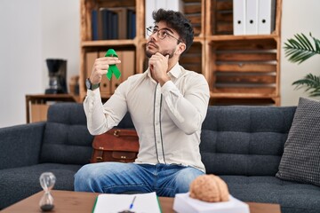 Obraz premium Hispanic man with beard holding support green ribbon at therapy clinic serious face thinking about question with hand on chin, thoughtful about confusing idea