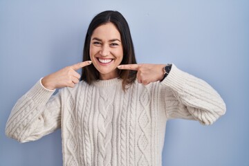 Young brunette woman standing over blue background smiling cheerful showing and pointing with fingers teeth and mouth. dental health concept.