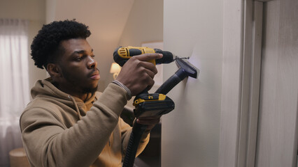 African American man in hoodie drills hole in concrete wall with hammer drill. Man does repairs in...