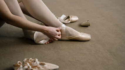 Ballerina sits on floor at dance studio and puts on pointe shoes before choreography lesson. Graceful ballet dancer prepares for performance or training. Classical ballet dance school. Feet close up.