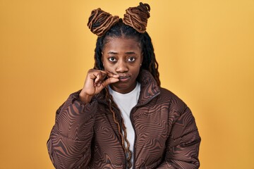 African woman with braided hair standing over yellow background mouth and lips shut as zip with...