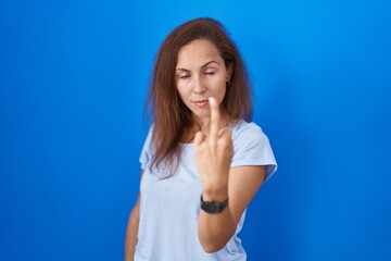 Brunette woman standing over blue background showing middle finger, impolite and rude fuck off expression