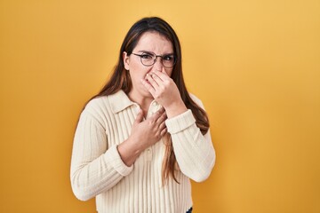 Young hispanic woman standing over yellow background smelling something stinky and disgusting, intolerable smell, holding breath with fingers on nose. bad smell