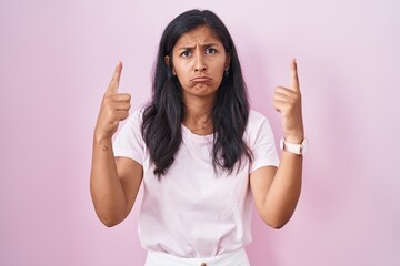 Young hispanic woman standing over pink background pointing up looking sad and upset, indicating direction with fingers, unhappy and depressed.