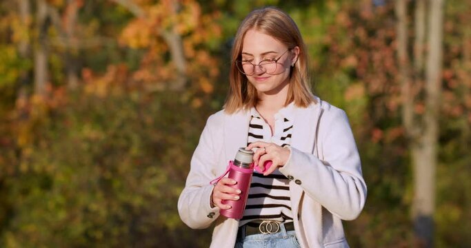 A beautiful girl in glasses opens a thermos with a hot drink and pours tea into a mug. Hot drinks to go outdoors in the park