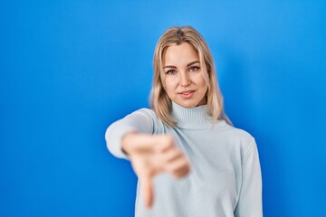 Fototapeta premium Young caucasian woman standing over blue background looking unhappy and angry showing rejection and negative with thumbs down gesture. bad expression.