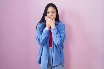 Young asian woman standing over pink background shocked covering mouth with hands for mistake....