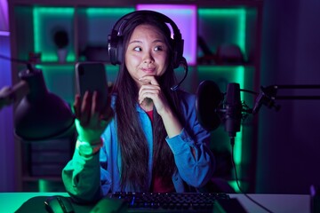 Young asian woman playing video games with smartphone with hand on chin thinking about question, pensive expression. smiling and thoughtful face. doubt concept.