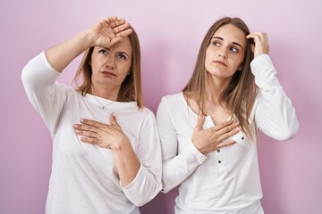 Middle age mother and young daughter standing over pink background touching forehead for illness and fever, flu and cold, virus sick