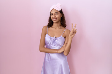 Young hispanic woman wearing sleep mask and nightgown smiling with happy face winking at the camera...