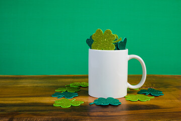 White mug full of clovers with glitter on wooden table. Green background.