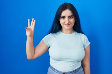 Young modern girl with blue hair standing over blue background showing and pointing up with fingers number three while smiling confident and happy.