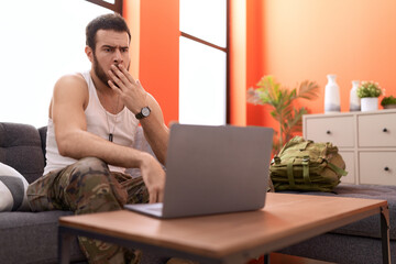 Young hispanic man wearing camouflage army uniform doing video call at home covering mouth with hand, shocked and afraid for mistake. surprised expression