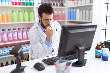 Young hispanic man pharmacist using computer with doubt expression at pharmacy