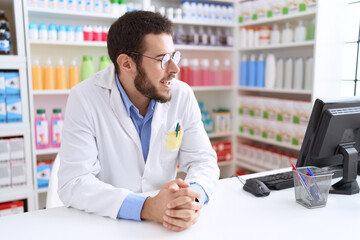Young hispanic man pharmacist smiling confident sitting on table at pharmacy