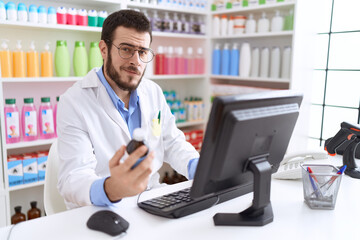 Young hispanic man pharmacist using computer holding medication bottle at pharmacy