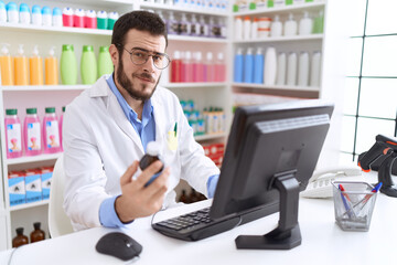 Fototapeta premium Young hispanic man pharmacist using computer holding medication bottle at pharmacy