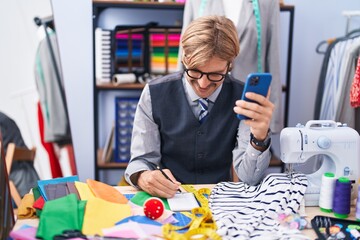 Young blond man tailor using smartphone writing on notebook at clothing factory