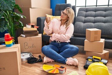 Young hispanic woman moving to a new home sitting on the floor looking proud, smiling doing thumbs up gesture to the side