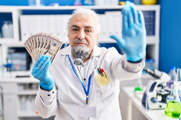 Middle age man with grey hair working at scientist laboratory holding money with open hand doing stop sign with serious and confident expression, defense gesture