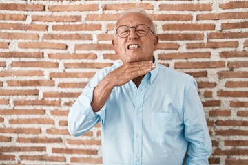 Senior man with grey hair standing over bricks wall cutting throat with hand as knife, threaten aggression with furious violence