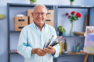 Senior grey-haired man artist smiling confident holding paintbrushes at art studio